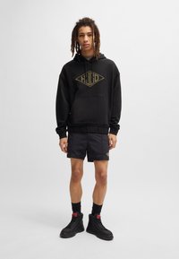 Felpa nera con logo dorato "HUGO", abbinata a pantaloni corti neri e stivali alti neri. Tessuto liscio, vestibilità casual, con dettagli delle calze.