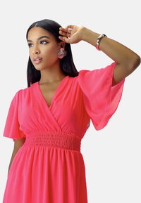 Robe rose fluide avec un col en V, taille froncée et manches volantées. Le mannequin porte des boucles d'oreilles fleuries et un bracelet métallique lisse.