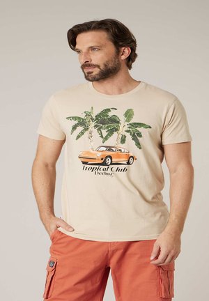 Homme barbu portant un t-shirt beige avec une voiture vintage orange et des palmiers verts, associé à un short cargo orange.