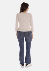 Maglietta lunga beige chiaro, dal design aderente. Abbinata a jeans blu a zampa d'elefante con tasche posteriori. Modella che indossa sneakers bianche.