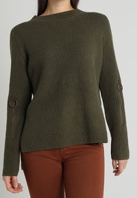 Frau in einem olivgrünen, gerippten Pullover mit Reißverschlussärmeln und rostfarbenen Hosen vor einem schlichten Hintergrund.