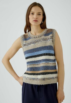 Frau mit langen braunen Haaren trägt einen ärmellosen Strickpullover mit blauen, schwarzen, weißen und beigen horizontalen Streifen und steht mit einer Hand in der Hüfte.