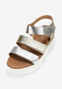 Sandalias metálicas con tres correas ajustables en plata y oro, que presentan un acabado texturizado y una suela de cuña de color claro.