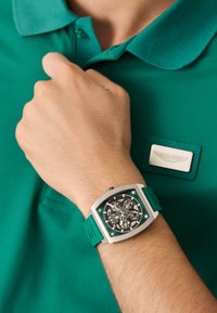 Kvadratiska pulksteņa ar caurspīdīgu seju un tirkīza akcentiem, kas saskaņota ar tirkīza gumijas jostu, valkāta uz rokas pie zaļas polo krekla.