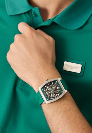 Ruutu kell läbipaistva näoga ja teal tooni aktsentidega, koos sobiva teal tooni kummirihmaga, kantud käel rohelise polo särgi vastu.