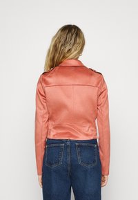 Korálová bunda z umělé kůže s přiléhavým střihem, klopovým límcem a dekorativními akcenty na ramenou. Natažená k modrým džínům.