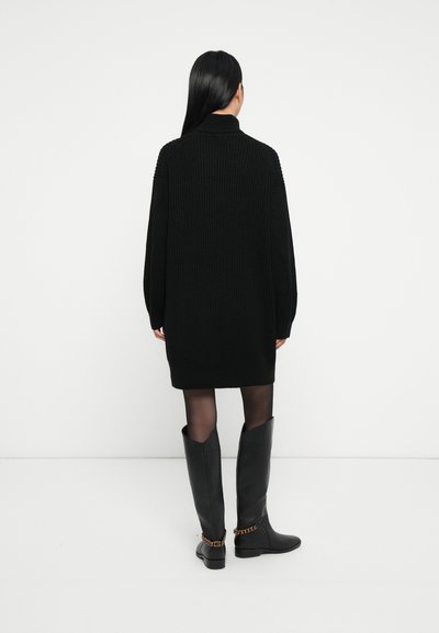 Femme aux longs cheveux noirs portant une robe pull noire en côtes, des collants noirs transparents et des bottes noires montantes avec des détails en chaîne dorée.