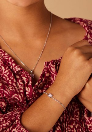 Zilveren ketting met een bloemenhanger en een bijpassende armband, voorzien van roze edelstenen. De achtergrond is een textiel met een gestructureerde stof in bordeauxrood.
