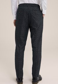 Osoba v tmavých vzorovaných kalhotách, černých botách a bílé košili s dlouhým rukávem, se stojí s rukama v kapsách na jednobarevném pozadí.