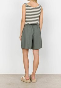 Žena v šedozelených volných šortkách, pruhovaném tílku bez rukávů a béžových podpatkových sandálech, stojící čelem ke bílé zdi.