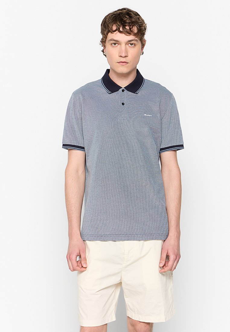 Jonge man in een grijze polo met korte mouwen en een donkere kraag, gekleed in beige shorts, staand tegen een effen witte achtergrond.