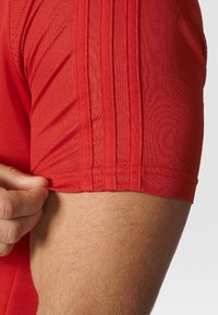 Primer plano del brazo de una persona que lleva una camiseta roja de manga corta con tres rayas verticales en la manga, la mano tirando ligeramente de la tela.