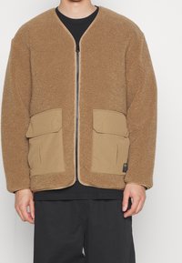 Giacca in pile marrone con zip frontale, due grandi tasche anteriori e un taglio ampio. Presenta un bordo in tessuto a contrasto lungo i bordi.