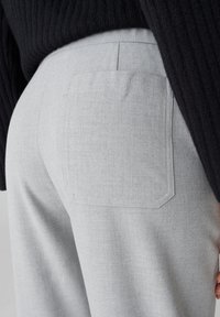 Pantalon gris clair en tissu texturé, avec une poche arrière unique et une coupe ajustée, associé à un pull noir à côtes.