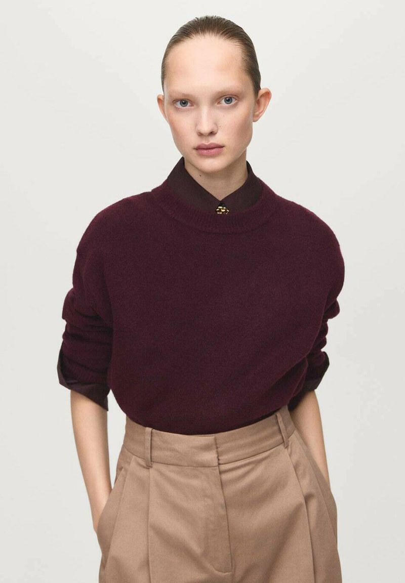 Maglione oversized bordeaux in maglia con colletto alto, dotato di maniche lunghe arrotolate. Abbinato a pantaloni beige a vita alta. Semplice dettaglio dei bottoni.