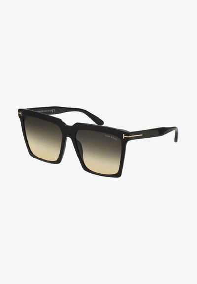 Gafas de sol negras cuadradas con un marco grueso y lentes degradados, que presentan un acabado en dorado y un diseño moderno. Logo de la marca visible en la patilla.