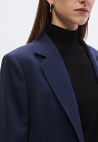 Granatowy blezer z dopasowanymi klapami, usztywnionymi ramionami i gładką fakturą, noszony na czarnym golfie. Widoczna srebrna kolczyk kołowy.