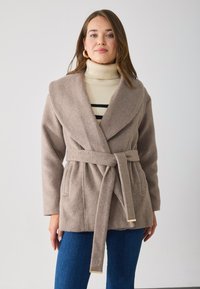Cappotto in misto lana beige con colletto a scialle, vita con cintura e due tasche frontali. Abbinato a un dolcevita a righe crema e denim.