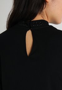 Top nero con una piccola apertura a chiave sul retro, decorato con borchie nere lungo il collo, realizzato in un materiale morbido.