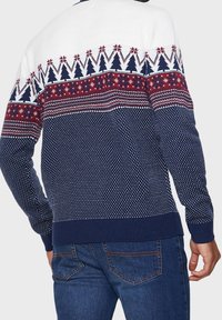 Pull en maille avec une section supérieure blanche à motifs, un corps bleu marine à pois, et des motifs texturés rouges et blancs sur la poitrine.