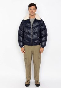 Man wearing navy blue puffer jacket with white shearling collar, gray shirt, khaki cargo pants, and tan hiking shoes standing against white background.