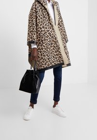Abrigo impermeable de estampado de leopardo con capucha, en tonos beige y marrón, combinado con un bolso tipo cubo negro, jeans oscuros y zapatillas blancas.