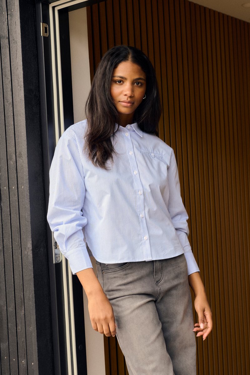 Femme aux longs cheveux foncés portant une chemise à rayures bleue claire boutonnée et un jean gris, se tenant dans un encadrement de porte avec un fond en panneaux de bois.