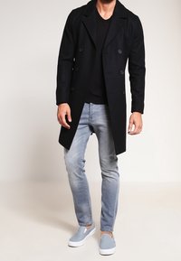 Abrigo negro de doble botonadura con solapas de muesca, llevado sobre una camisa negra y unos jeans azul claro. Zapatos grises tipo slip-on completan el conjunto.