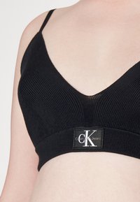Musta ribbed bralette, jossa on syvä V-pääntie ja ohuet hihnat. Sisältää brändätyn elastisen nauhan, jossa on "Calvin Klein Jeans" - logo.