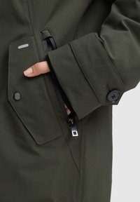 Grüne wasserdichte Jacke mit einer Reißverschlusstasche und Knopfdetails an den Ärmeln. Glatte Textur und modernes Design.
