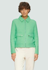 Veste verte coupée avec un col, fermeture  éclair devant et deux poches avant. Fabriquée en tissu texturé, elle présente une coupe décontractée et un design minimaliste.