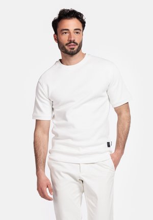 Weißes Crewneck-Sweatshirt mit kurzen Ärmeln, weicher Textur und sauberen Säumen. Kombiniert mit passenden, hellfarbigen Hosen, mit minimaler Markenkennzeichnung.