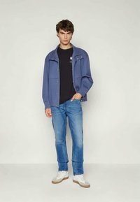 Plava traper jakna opuštenog kroja preko crne majice, u kombinaciji sa svijetloplavim trapericama i bijelim tenisicama, stojeći na neutranoj pozadini.