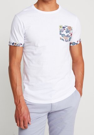 Mężczyzna ubrany w biały t-shirt z krótkim rękawem z kwiatowym nadrukiem na kieszeni i mankietach, w połączeniu z jasnoszarymi spodniami.