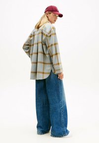 Chemise à carreaux bleue et beige, coupe oversize, associée à un jean large bleu clair. Casquette rouge foncé. Tissu doux, design décontracté.