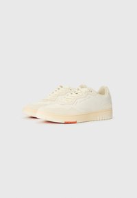 Cream-colored sneakers with a suede and smooth finish, featuring a rounded toe, lace-up design, and a soft, contrasting orange accent on the sole.