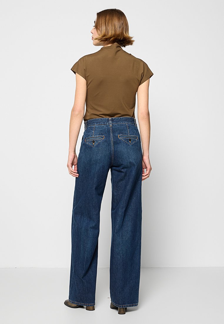 Brązowa koszula z krótkim rękawem zestawiona z niebieskimi dżinsami z wysokim stanem i szerokimi nogawkami, które mają tylne kieszenie i detaliczne przeszycia.