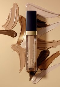 Flytande concealer i en genomskinlig flaska med svart kork, omgiven av olika nyanser av kräm, tan och brun makeup-prover på en beige bakgrund.