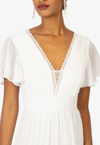 Witte jurk met korte, gerimpelde mouwen, een diepe V-halslijn afgezet met kant. Geplooide stof met een getailleerde taille en een vloeiende rok.