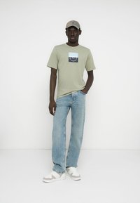 Ανοιχτό πράσινο βαμβακερό t-shirt με γραφικό σχέδιο, μπλε τζιν και λευκά αθλητικά παπούτσια με γκρι λεπτομέρειες. Το μοντέλο φοράει μπεζ καπέλο.