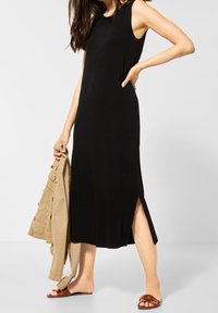 Kvinna som bär en ärmlös svart midiklänning med sidssprund, håller en beige jacka och har på sig bruna platta sandaler mot en enkel vit bakgrund.