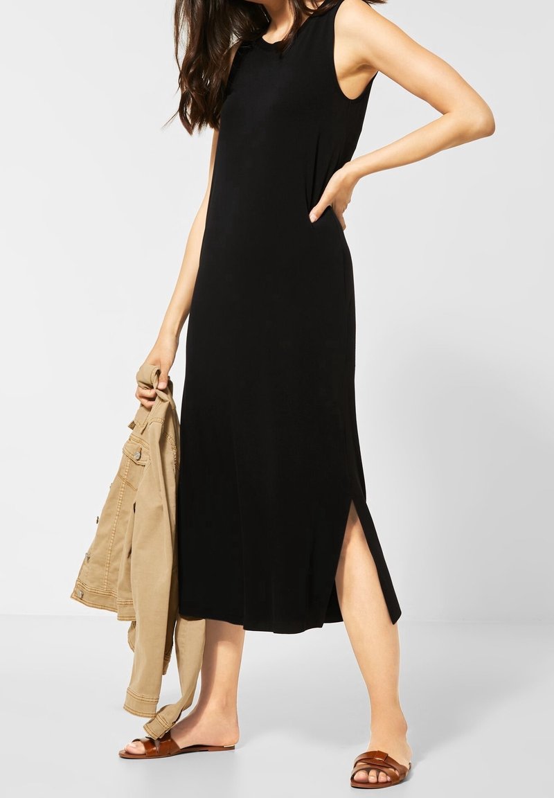 Kvinna som bär en ärmlös svart midiklänning med sidssprund, håller en beige jacka och har på sig bruna platta sandaler mot en enkel vit bakgrund.