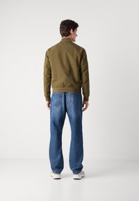 Olive green bomber jacket with ribbed cuffs and hem, paired with loose-fit blue jeans and light-colored sneakers, viewed from the back.
