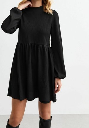 Woman wearing a black, long-sleeve, textured mini dress with a high neckline and gathered waist, paired with black knee-high boots.