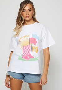 Camiseta blanca con un diseño gráfico que presenta botas de cuadros rosas, un sol, banderas coloridas y un símbolo de paz, todo sobre un fondo de hierba verde.