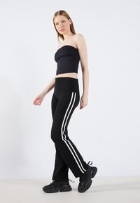 Crop top noir sans bretelles et pantalons noirs avec des rayures blanches sur les côtés, associés à des baskets noires volumineuses sur une surface claire.