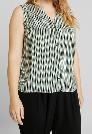 Bez rękawów bluzka na guziki z czarno-białym geometrycznym wzorem, noszona z czarnymi spodniami przez osobę o długich blond włosach.