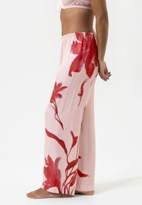 Femme portant un pantalon large rose clair à grands motifs floraux rouges, associé à un soutien-gorge en dentelle rose pâle, pieds nus sur un fond blanc.