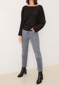 Femme portant un pull noir côtelé à manches longues, un jean gris droit et des bottines noires à talons, debout contre un mur blanc.