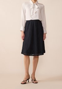 Valkoinen paita, jossa on kaulakoru, yhdistettynä mustaan pitsihameeseen, jossa on kukkakuviot ja aaltoileva helma, täydentävät mustat korkokengät.